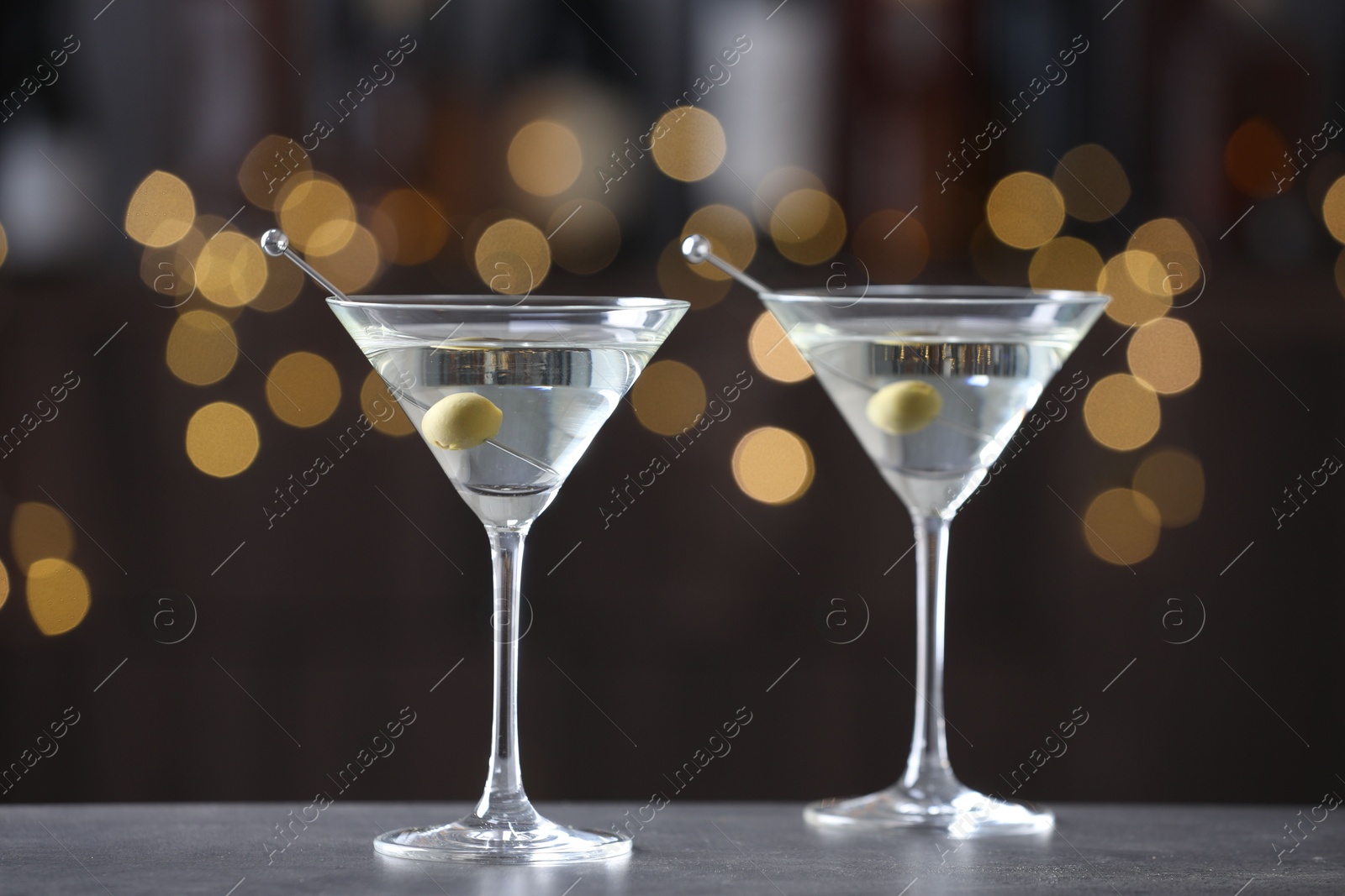 Photo of Martini cocktail with olives in glasses on grey table against blurred background. Bokeh effect