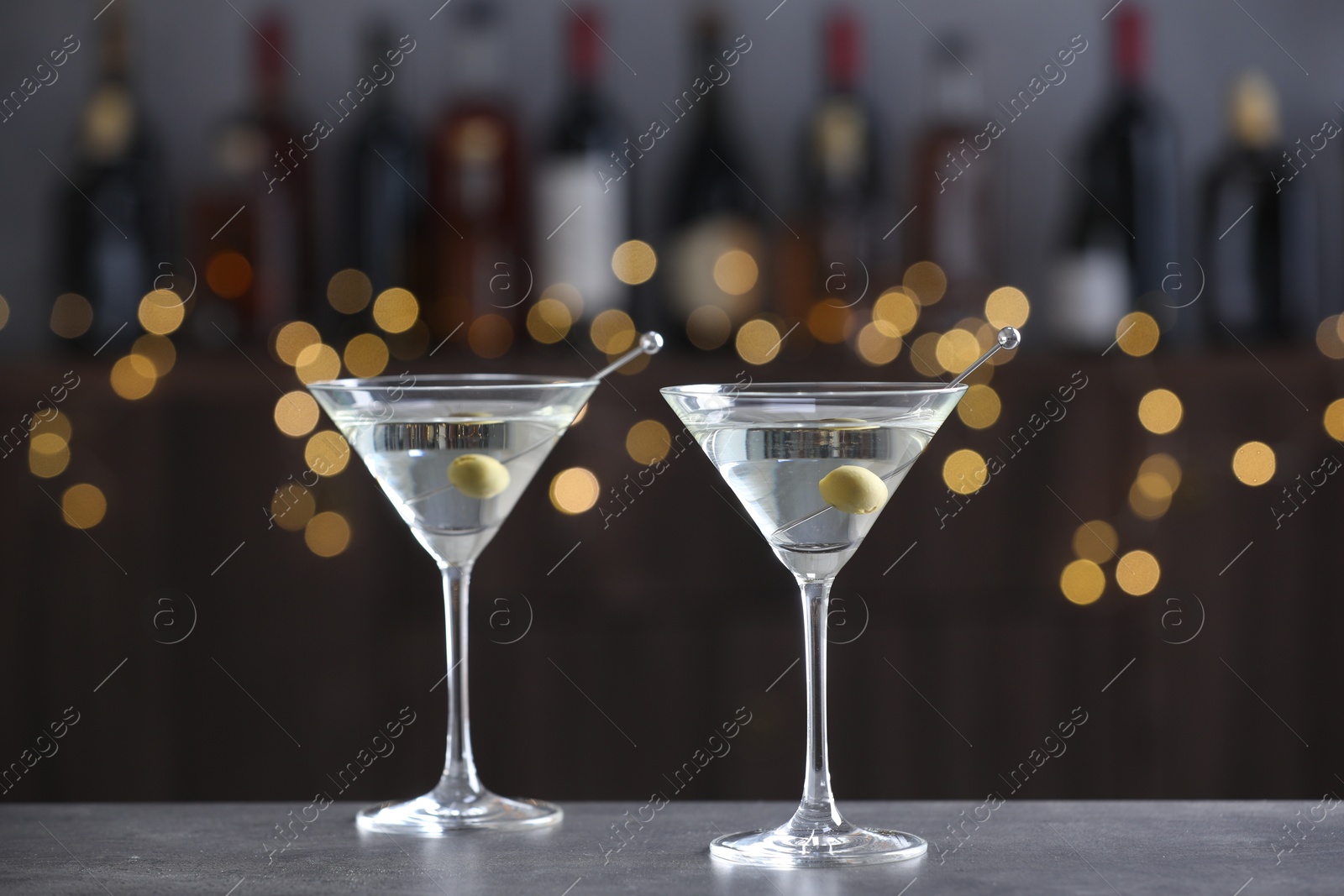 Photo of Martini cocktail with olives in glasses on grey table against blurred background. Bokeh effect