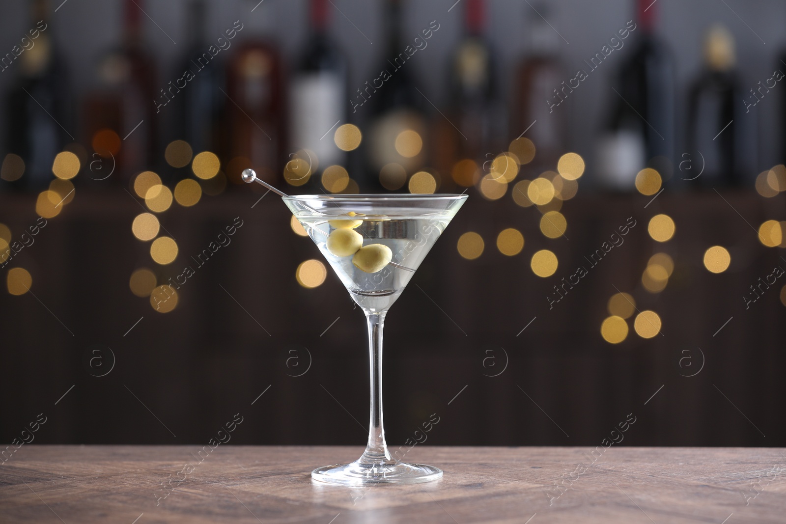 Photo of Martini cocktail with olives in glass on wooden table against blurred background. Bokeh effect