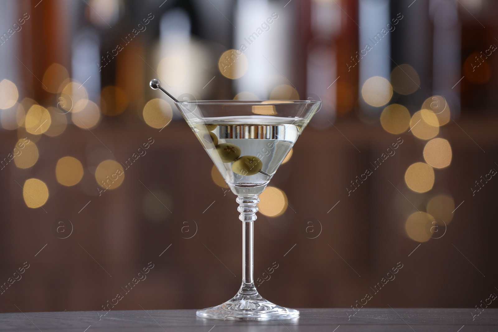 Photo of Martini cocktail with olives in glass on wooden table against blurred background. Bokeh effect