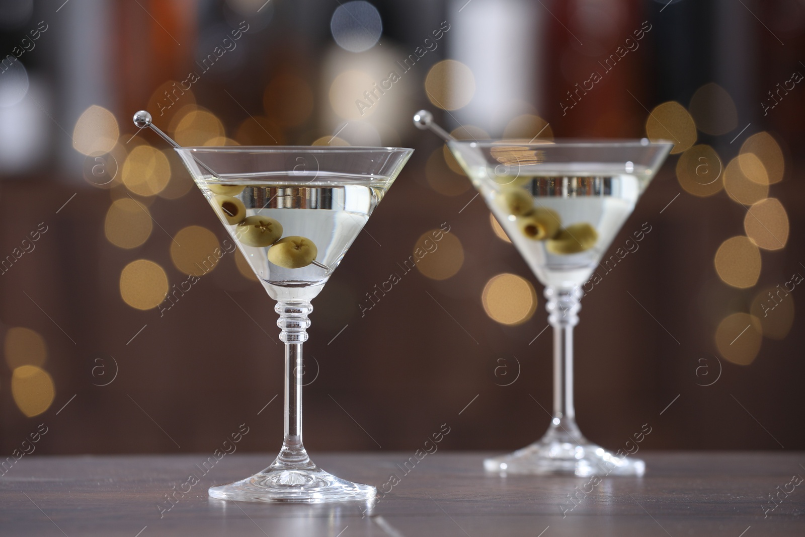 Photo of Martini cocktail with olives in glasses on wooden table against blurred background. Bokeh effect