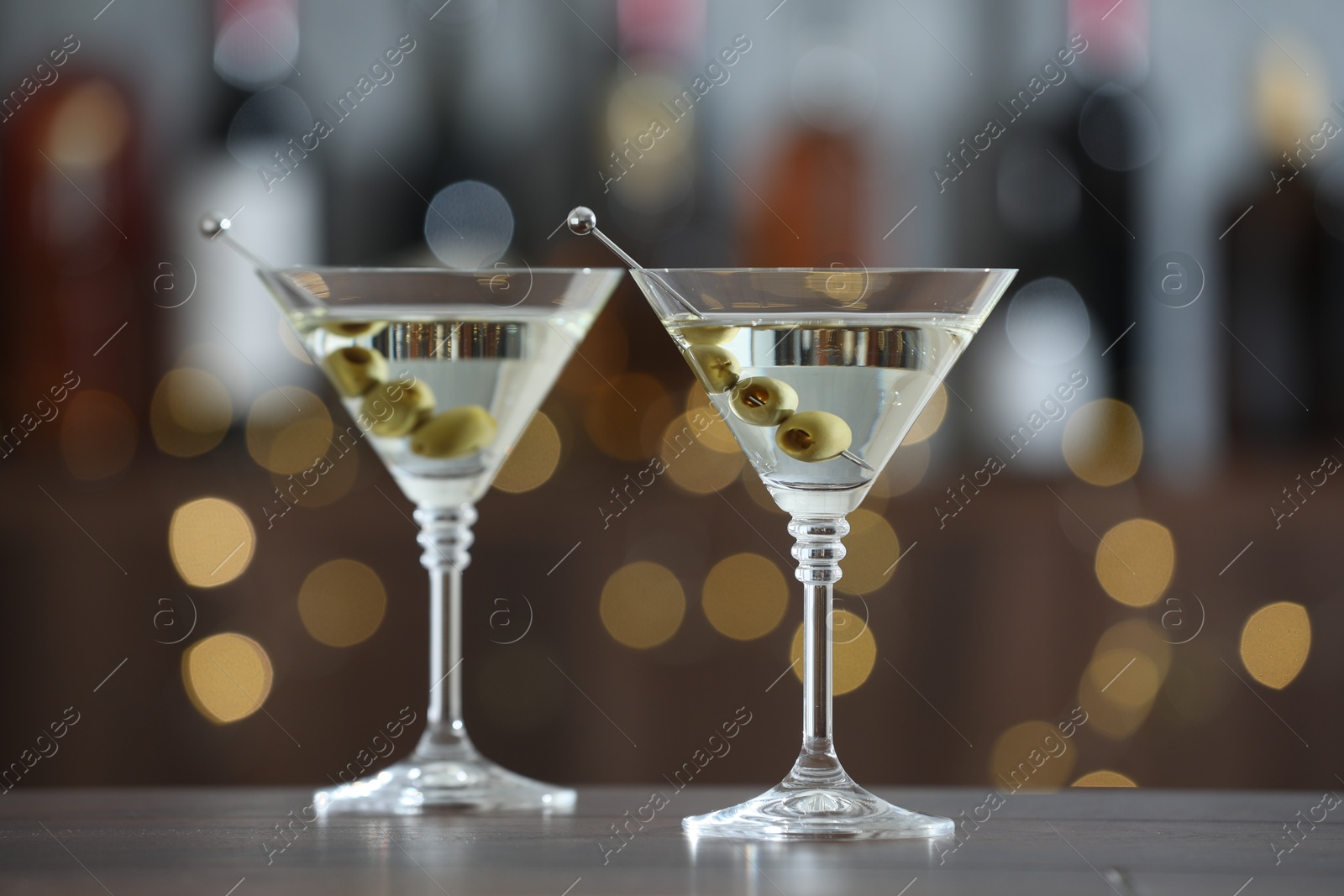 Photo of Martini cocktail with olives in glasses on wooden table against blurred background. Bokeh effect