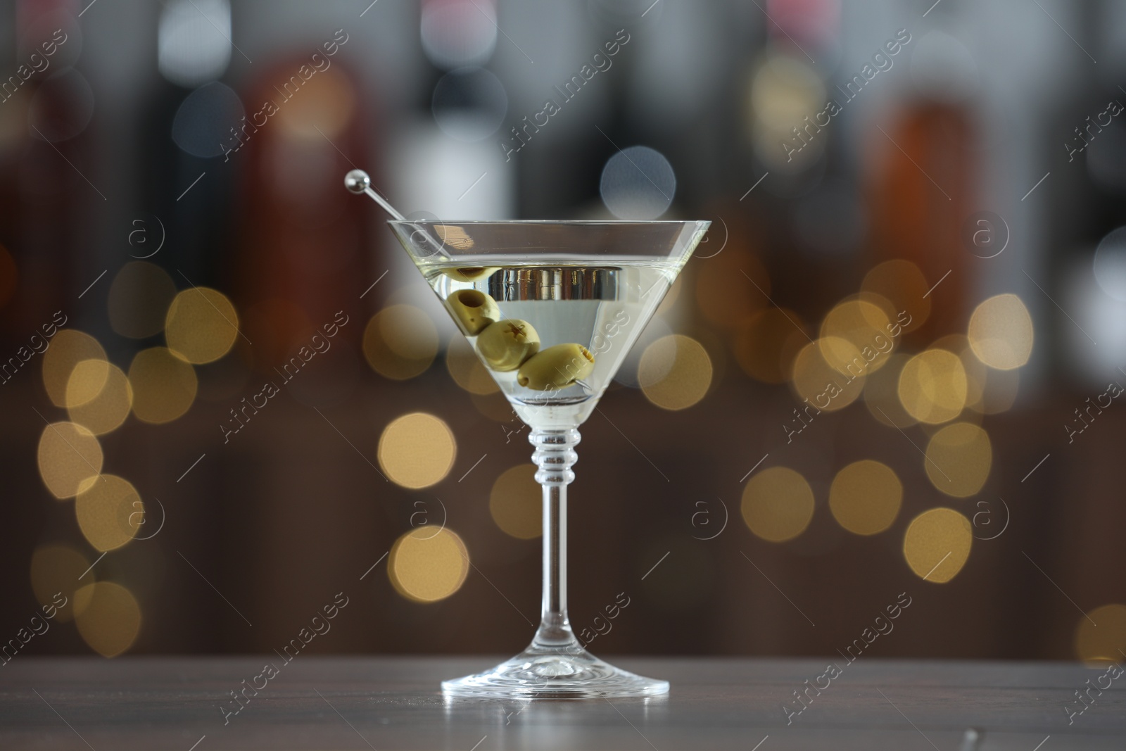 Photo of Martini cocktail with olives in glass on wooden table against blurred background. Bokeh effect