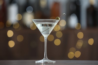 Photo of Martini cocktail with olive in glass on wooden table against blurred background. Bokeh effect