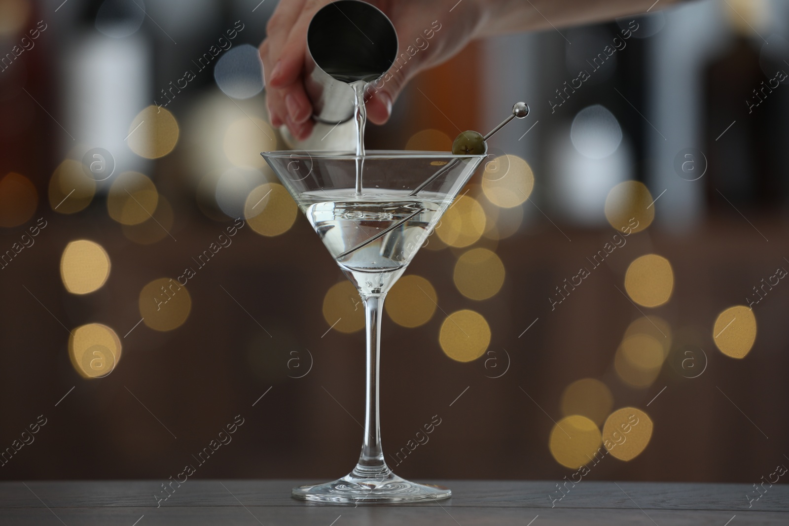 Photo of Woman making tasty martini cocktail with olives at wooden table on blurred background, closeup. Bokeh effect