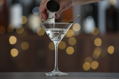 Photo of Woman making tasty martini cocktail with olives at wooden table on blurred background, closeup. Bokeh effect