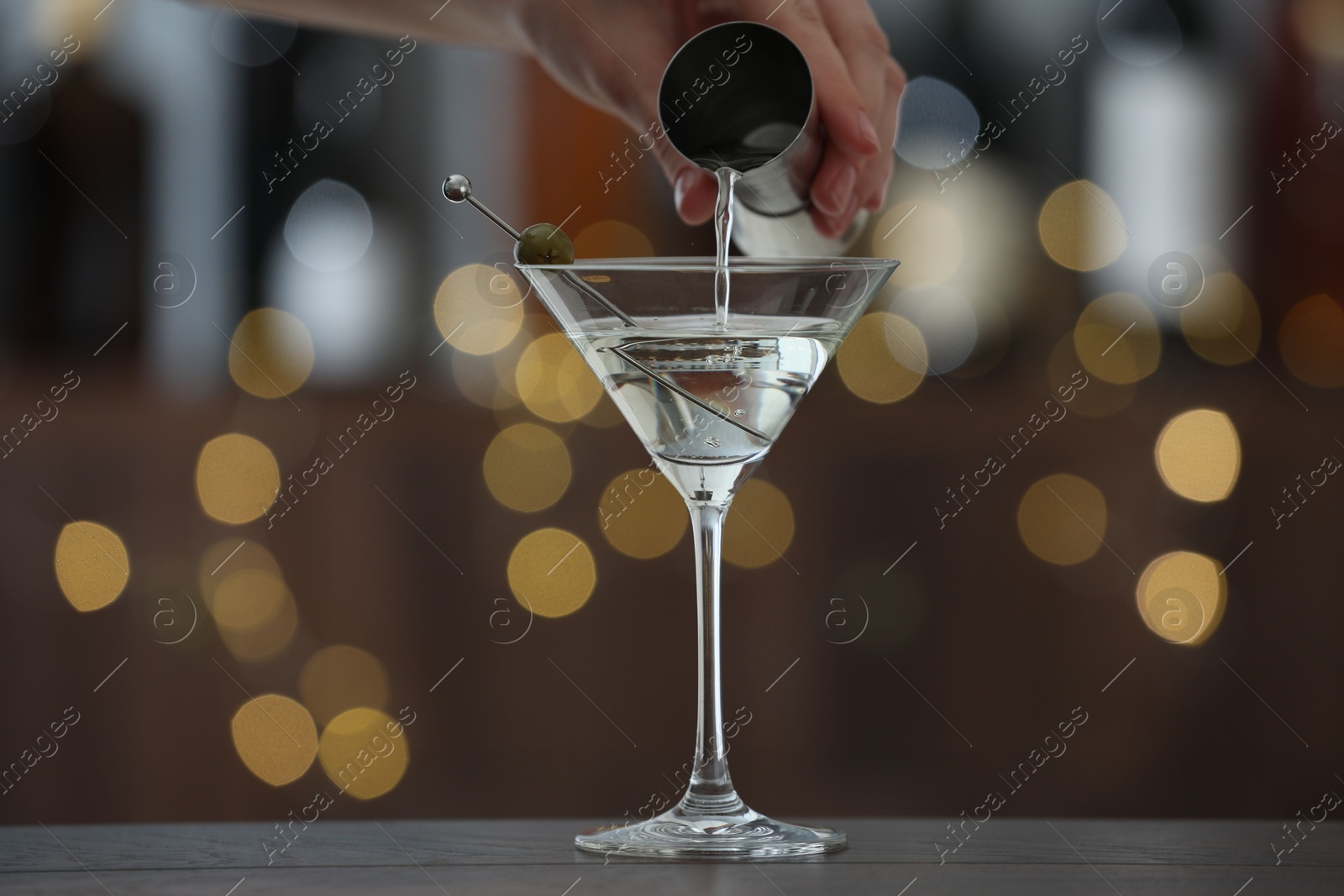 Photo of Woman making tasty martini cocktail with olives at wooden table on blurred background, closeup. Bokeh effect