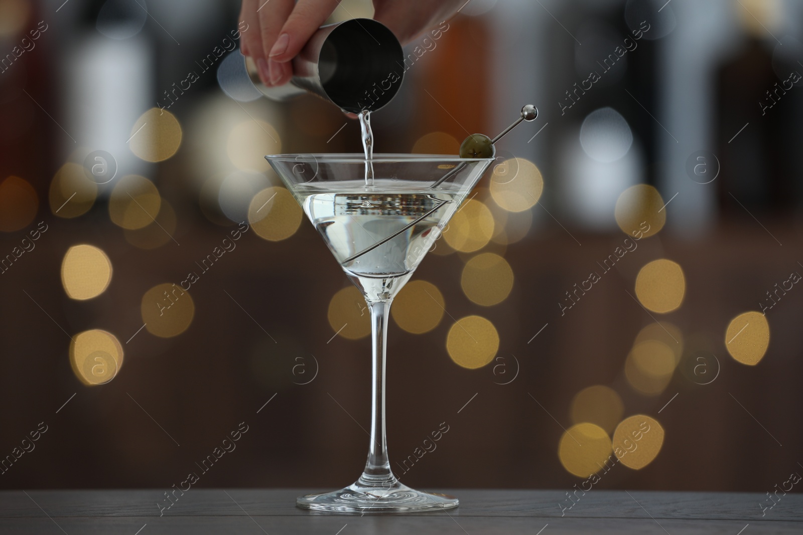 Photo of Woman making tasty martini cocktail with olives at wooden table on blurred background, closeup. Bokeh effect