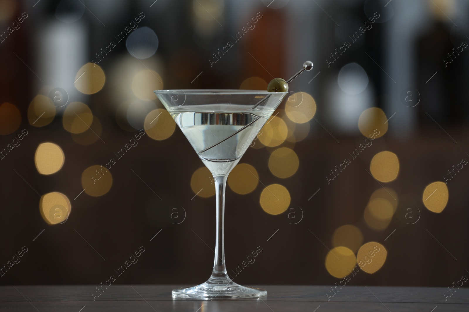 Photo of Martini cocktail with olive in glass on wooden table against blurred background. Bokeh effect