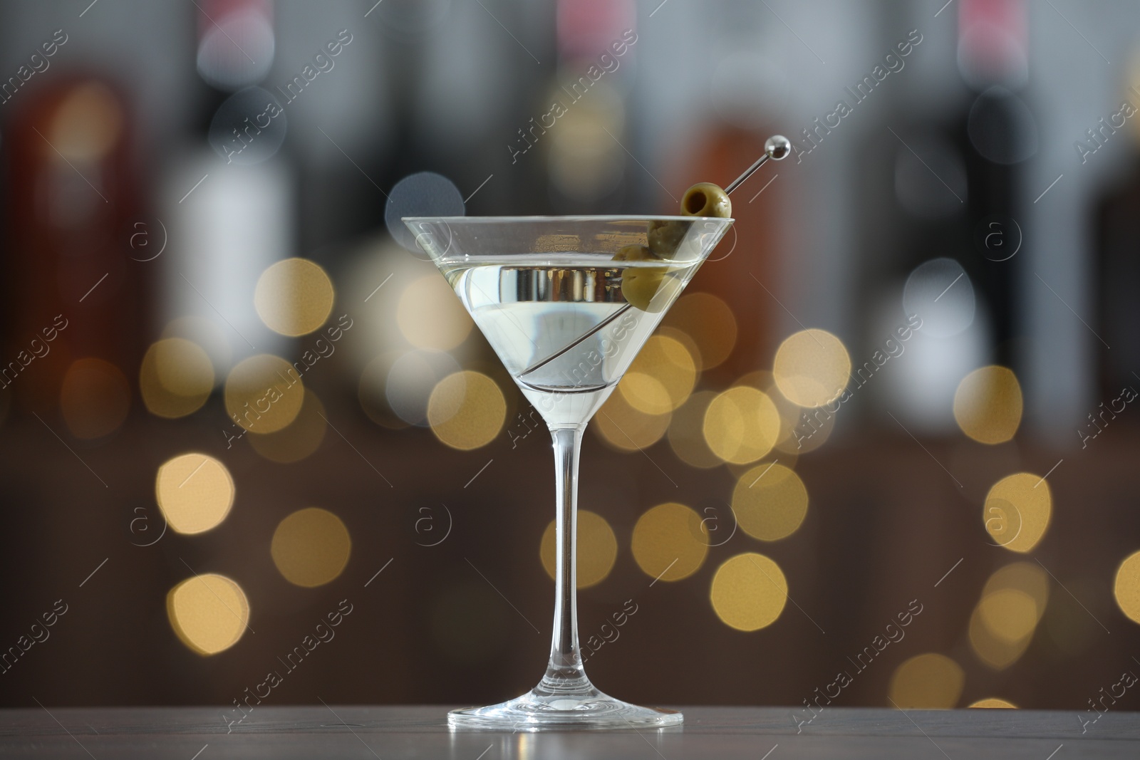 Photo of Martini cocktail with olives in glass on wooden table against blurred background. Bokeh effect