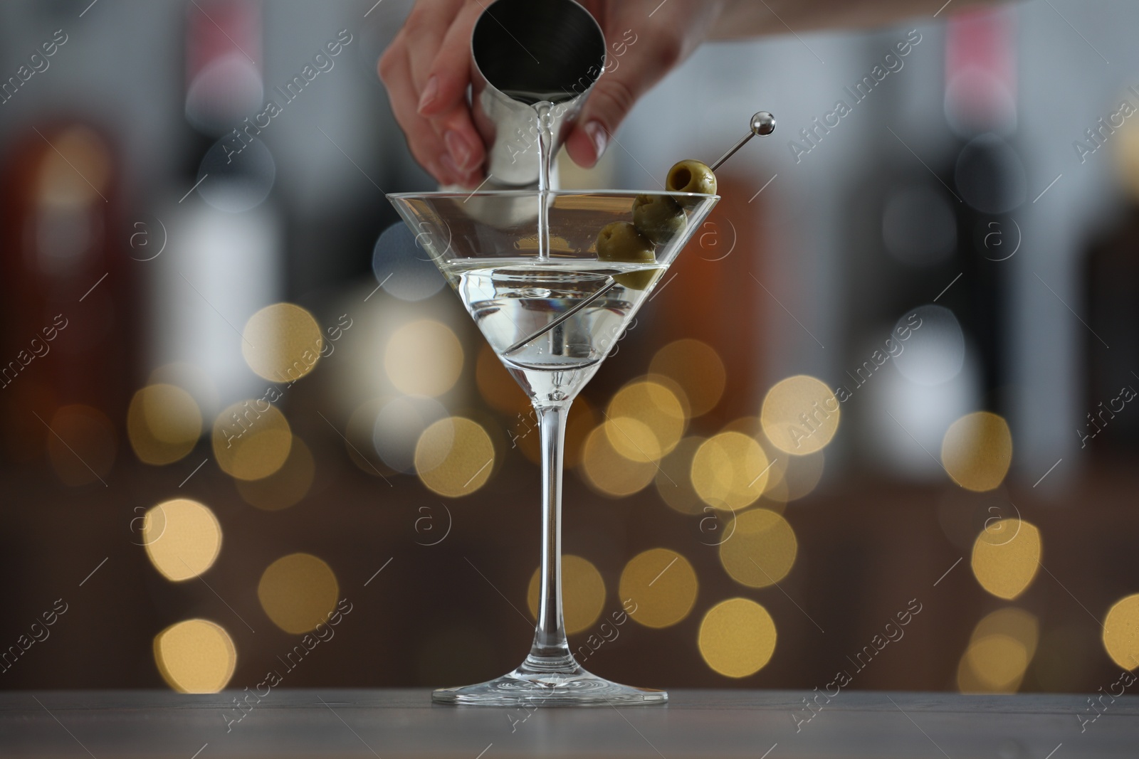 Photo of Woman making tasty martini cocktail with olives at wooden table on blurred background, closeup. Bokeh effect