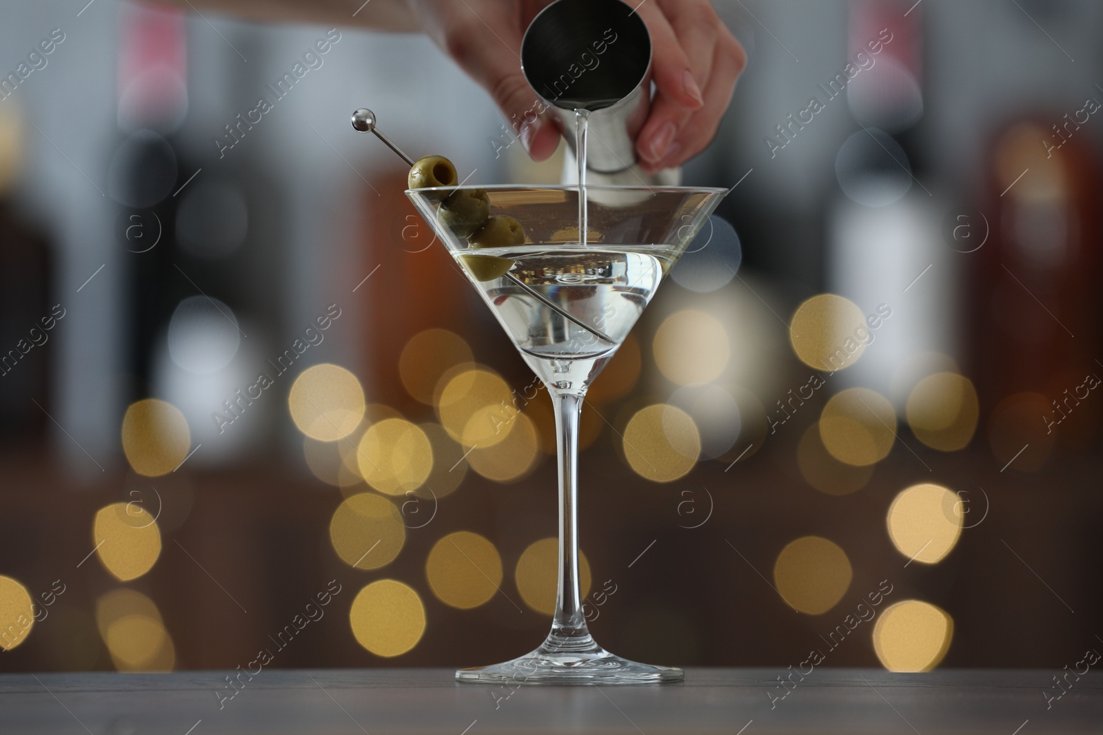 Photo of Woman making tasty martini cocktail with olives at wooden table on blurred background, closeup. Bokeh effect