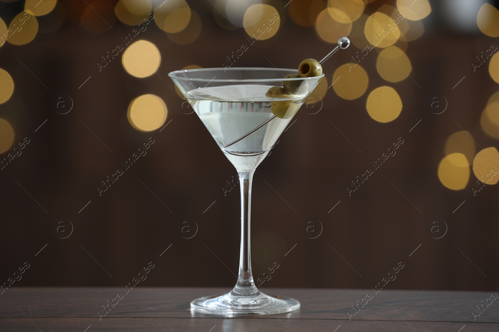 Photo of Martini cocktail with olives in glass on wooden table against blurred background. Bokeh effect