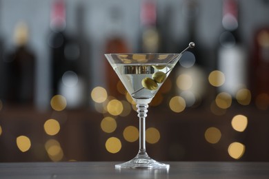 Photo of Martini cocktail with olives in glass on wooden table against blurred background. Bokeh effect