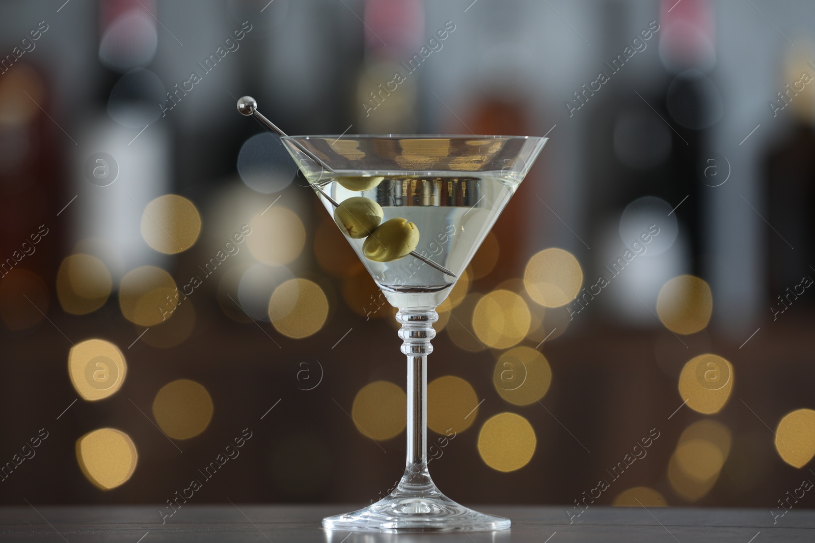 Photo of Martini cocktail with olives in glass on wooden table against blurred background. Bokeh effect