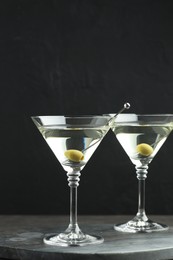 Photo of Martini cocktail with olives in glasses on grey table against black background