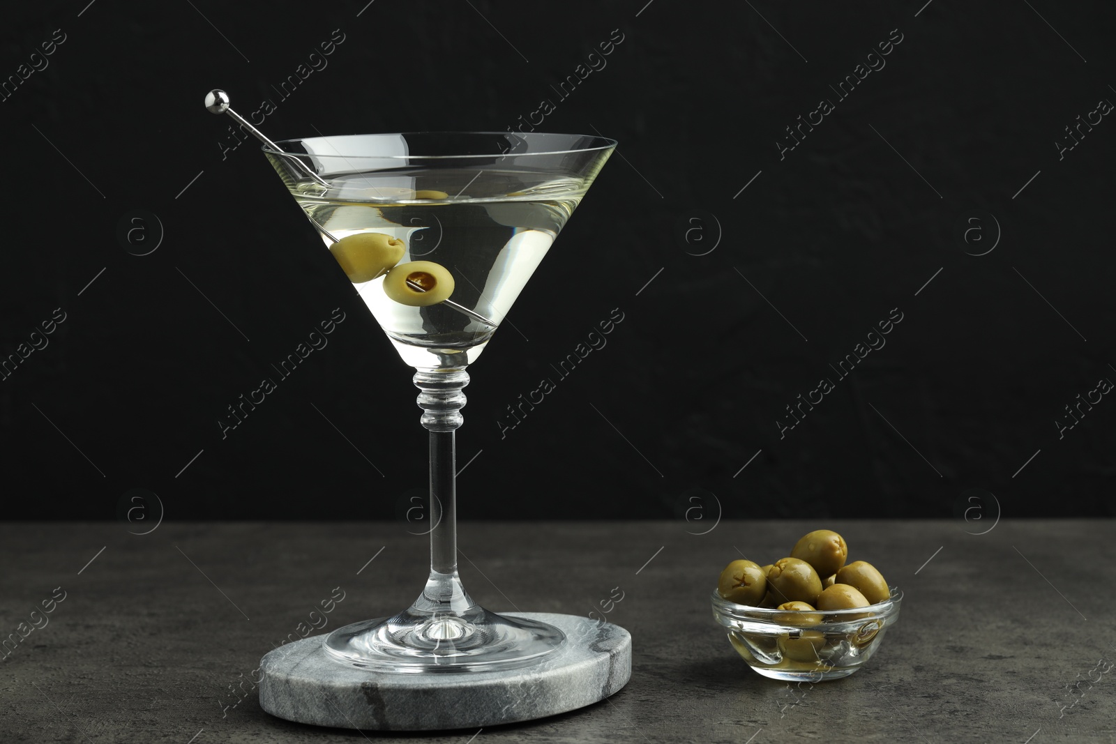 Photo of Martini cocktail with olives in glass on grey table against black background