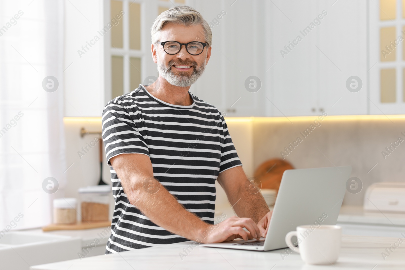 Photo of Happy middle aged man using laptop at white marble table in kitchen