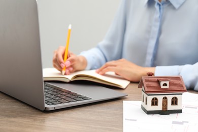 Photo of House hunting. Woman with laptop, papers, notebook and house figure at wooden table, closeup