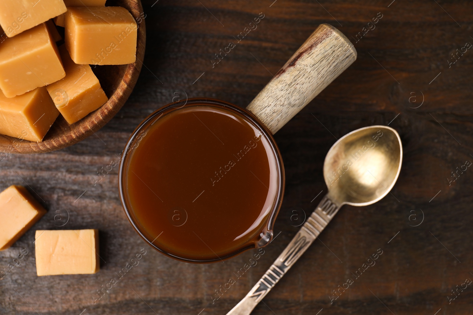 Photo of Tasty caramel sauce in glass measuring cup, sweet candies and spoon on wooden table, flat lay