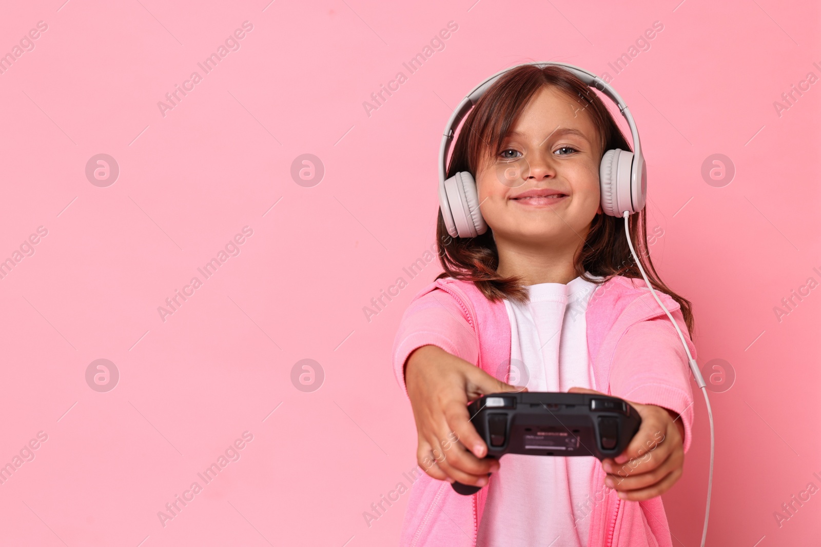 Photo of Cute little girl in headphones playing video game with controller on pink background. Space for text