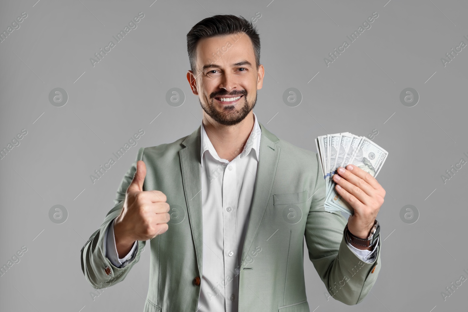 Photo of Portrait of smiling banker with dollar banknotes showing thumbs up on grey background