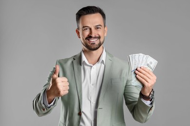 Photo of Portrait of smiling banker with dollar banknotes showing thumbs up on grey background