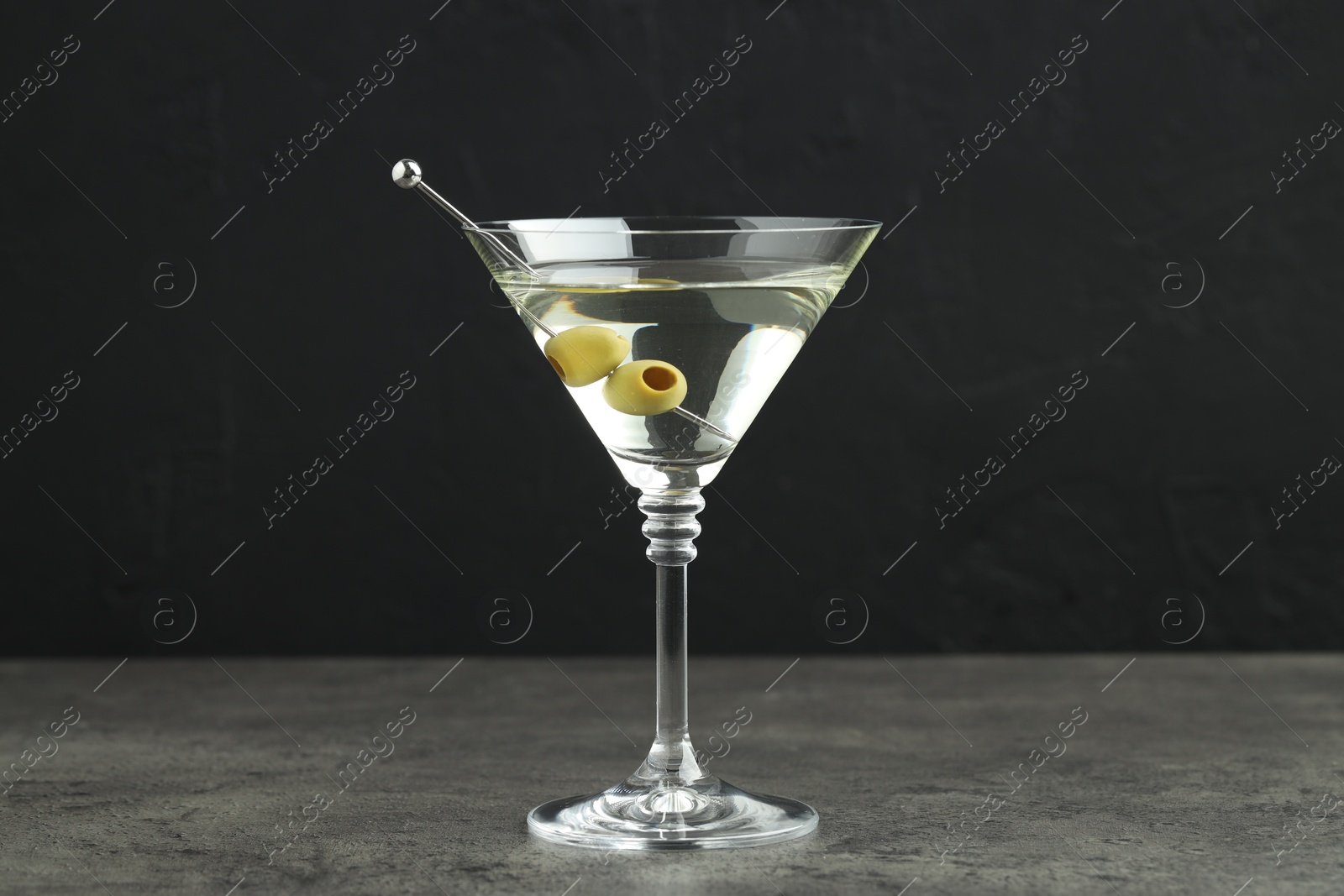 Photo of Martini cocktail with olives in glass on grey table against black background