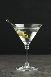 Photo of Martini cocktail with olives in glass on grey table against black background