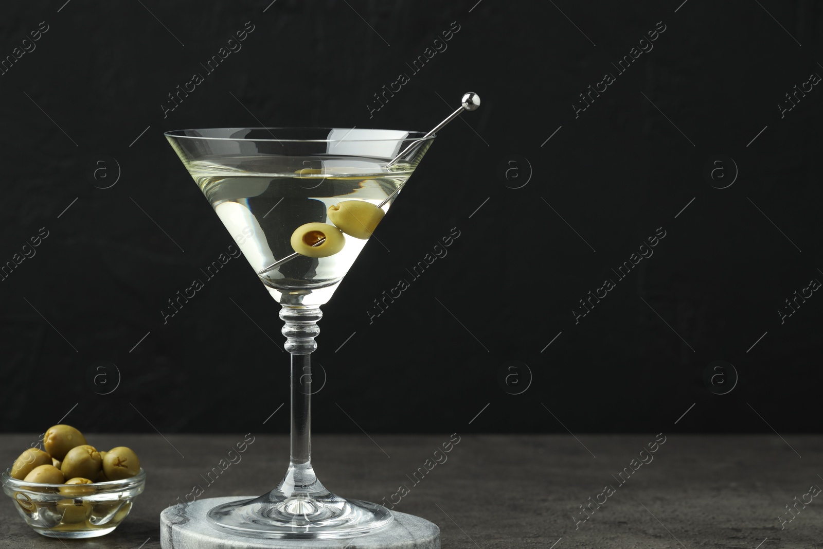 Photo of Martini cocktail with olives in glass on grey table against black background, space for text