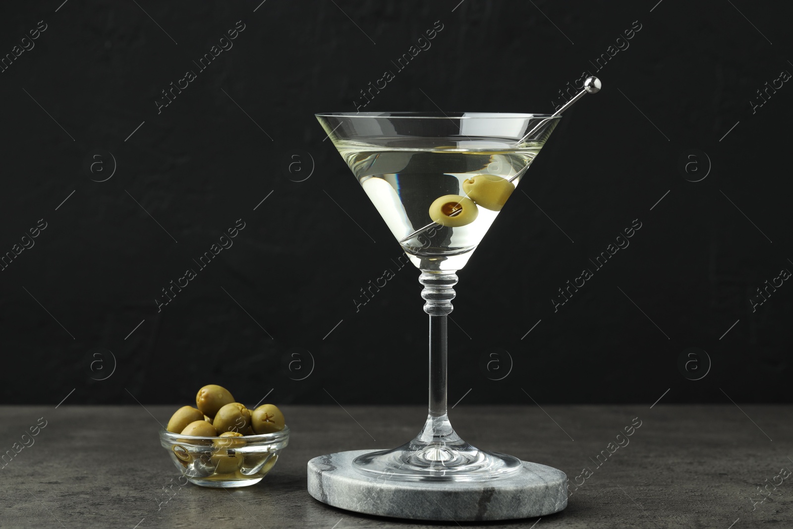 Photo of Martini cocktail with olives in glass on grey table against black background