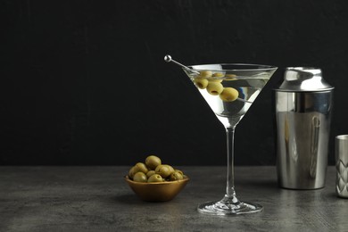 Photo of Martini cocktail with olives in glass and shaker on grey table against black background, space for text