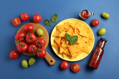 Photo of Tasty ketchup, nachos, tomatoes and basil on blue background, flat lay