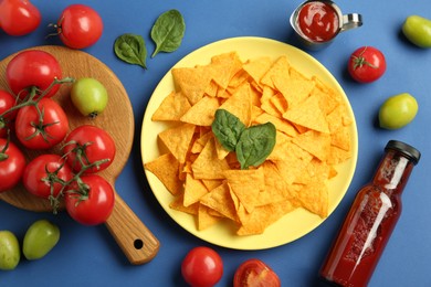 Photo of Tasty ketchup, nachos, tomatoes and basil on blue background, flat lay