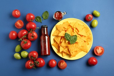 Photo of Tasty ketchup, nachos, tomatoes and basil on blue background, flat lay