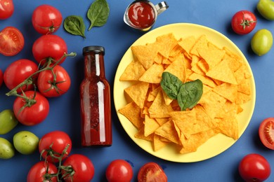 Photo of Tasty ketchup, nachos, tomatoes and basil on blue background, flat lay