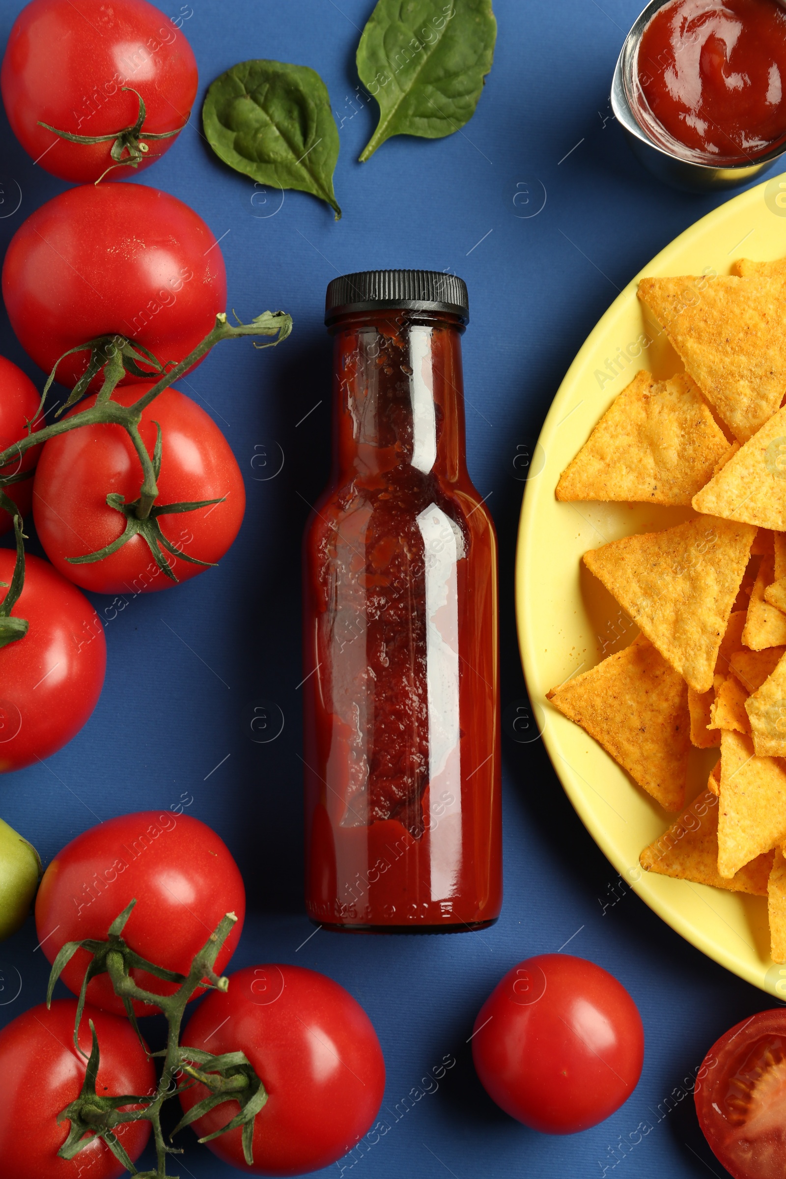 Photo of Tasty ketchup, nachos, tomatoes and basil on blue background, flat lay