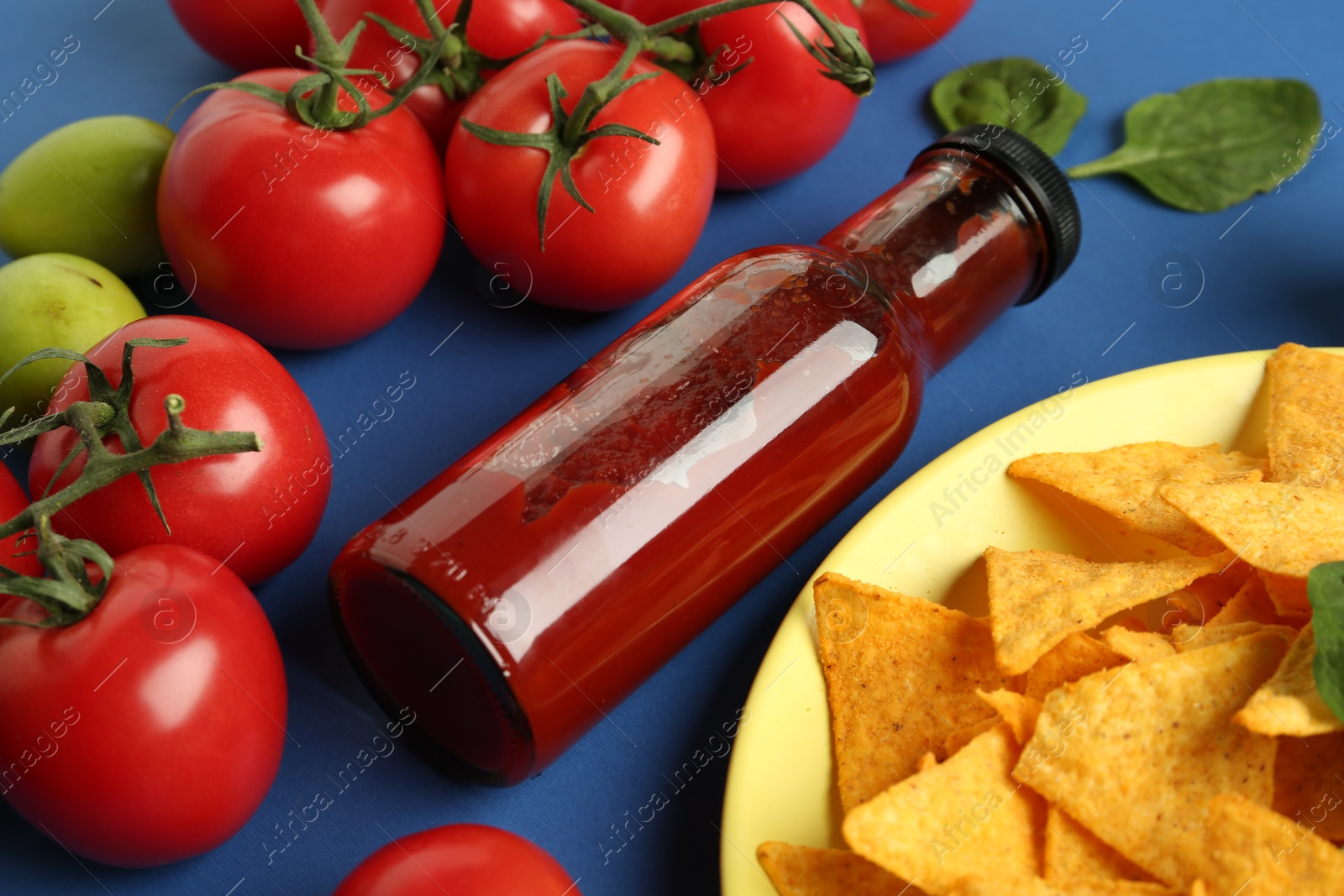 Photo of Tasty ketchup, nachos, tomatoes and basil on blue background, closeup