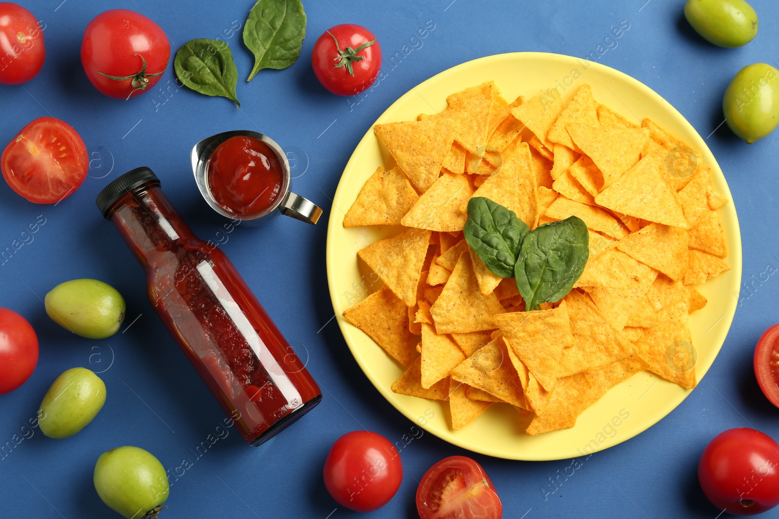 Photo of Tasty ketchup, nachos, tomatoes and basil on blue background, flat lay