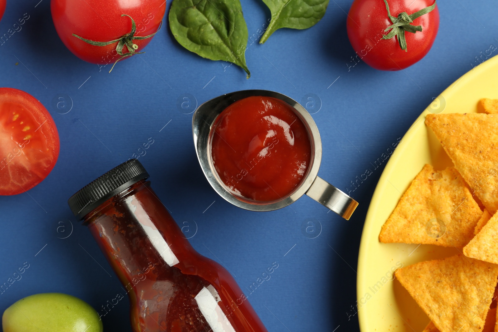 Photo of Tasty ketchup, nachos, tomatoes and basil on blue background, flat lay