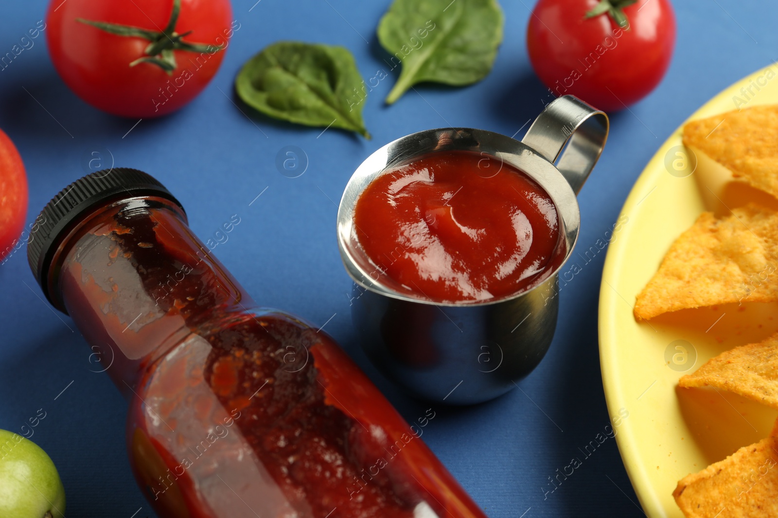 Photo of Tasty ketchup, nachos, tomatoes and basil on blue background, closeup