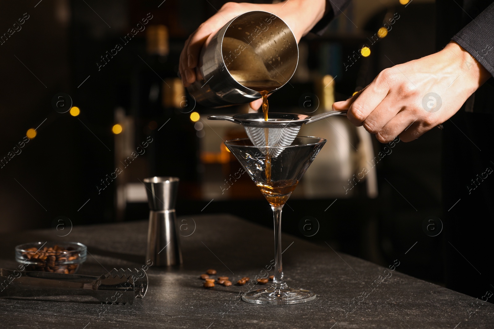 Photo of Bartender making delicious espresso martini at dark table against blurred lights, closeup