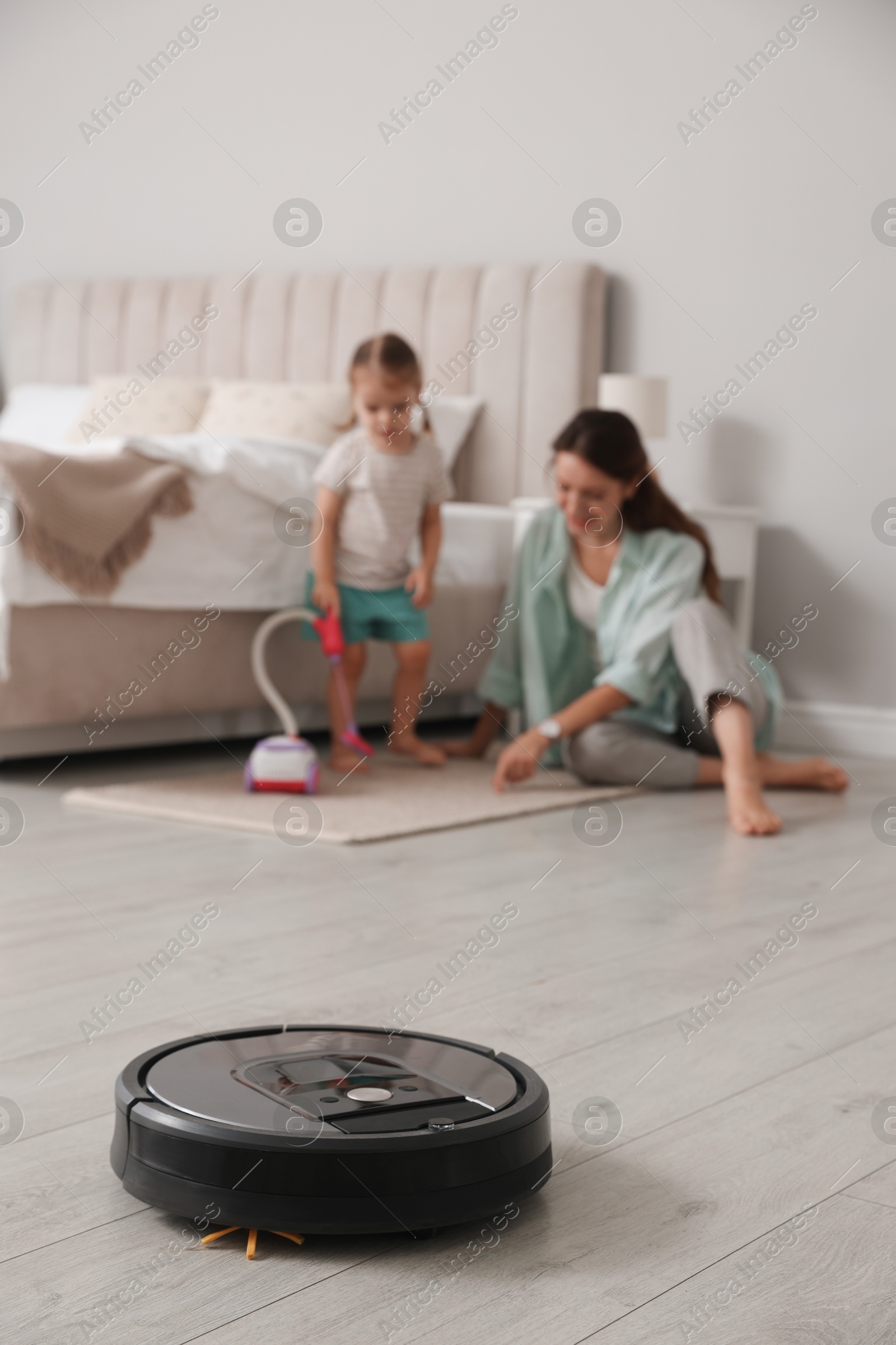 Photo of Mother spending time with her daughter in bedroom, focus on robotic vacuum cleaner