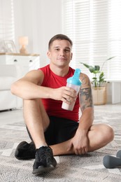 Photo of Athletic man with shaker of protein drink sitting on carpet at home