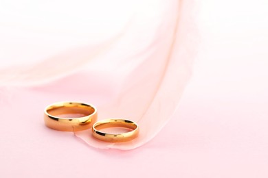 Photo of Beautiful golden wedding rings and feather on pink background, closeup