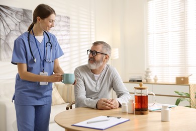 Photo of Caregiver giving cup of drink to senior man indoors. Home health care service