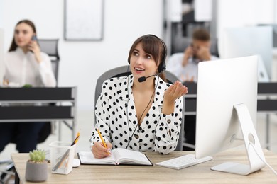 Photo of Saleswoman making notes while talking to client via headset at desk in office