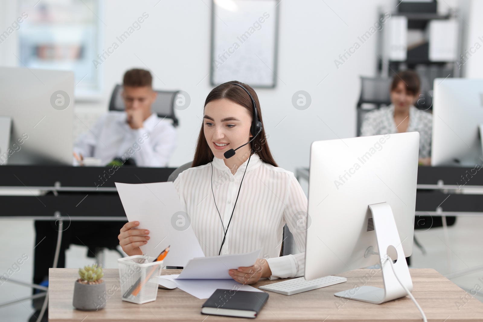 Photo of Saleswoman checking documents while talking to client via headset at desk in office