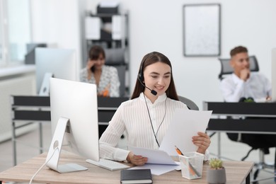 Photo of Saleswoman checking documents while talking to client via headset at desk in office