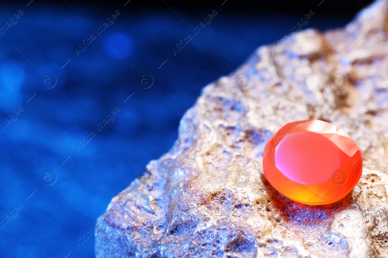 Photo of Beautiful shiny orange gemstone on stone against blue background, space for text